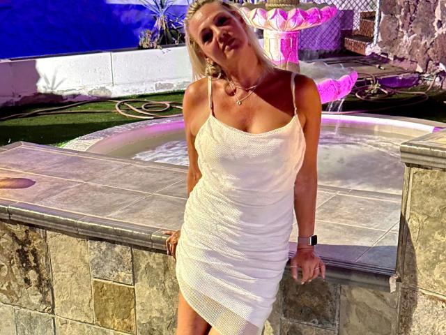 Blueberry-10-hot, blonde, wearing a tight white dress, showing her ass, leaning on a stone wall, wristwatch, pink necklace, night, pool, purple lights.