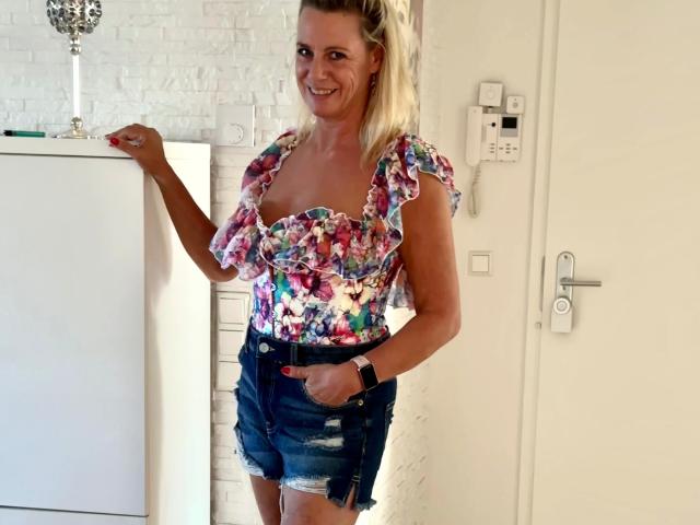 Blueberry-10-hot with blonde hair, smiling, wearing a colorful floral top with puffy sleeves and denim shorts, hand in pocket, standing by a white door, wearing a black wristband.