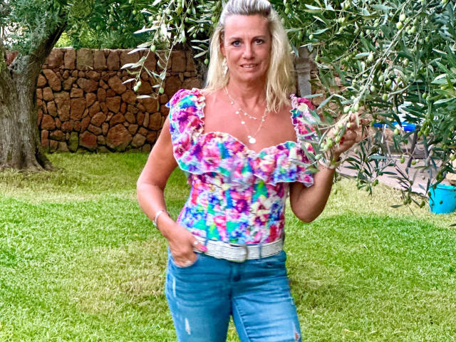 Blueberry-10-hot, blonde woman with medium tits, wearing a colorful floral top, blue jeans, and a white belt, holding olive branches, smiling outdoors, green grass, stone wall.