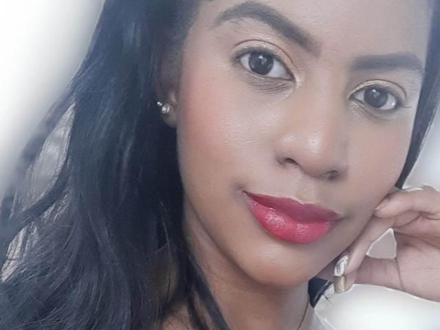 KelleySinedd has dark skin, black hair, red lipstick, and brown eyes with subtle makeup. Her right hand rests near her face, showing a diamond earring.