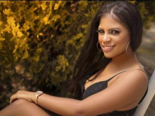 KelleySinedd, brown-skinned, smiling with long dark hair, wearing a black dress, hoop earrings, and a wristwatch, sitting outdoors, showcasing her small tits and curvy ass.