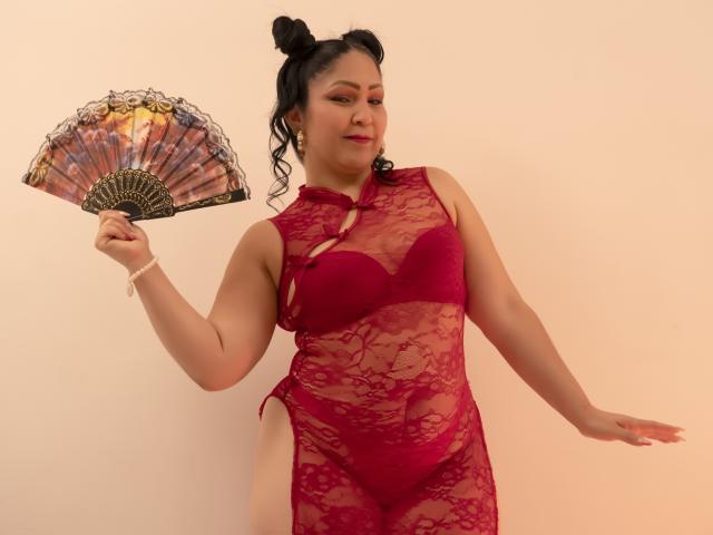 ArabelaFoxy, con dos moños negros, vistiendo lencería roja de encaje, sosteniendo un abanico, tiene senos de tamaño medio y un cuerpo curvilíneo y sexy, de pie frente a un fondo liso..