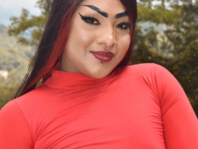 AuroraRossx with red shirt, thick black eyebrows, black eyeliner, red lipstick, nose ring, lip piercing, and red-tipped black hair, smiling confidently. Boobs not visible.