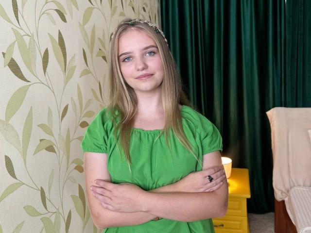 LunaBellaHottiest with blonde hair, green eyes, and small tits, wearing a green dress, arms crossed, standing in front of green curtains, and a yellow lamp on a nightstand.