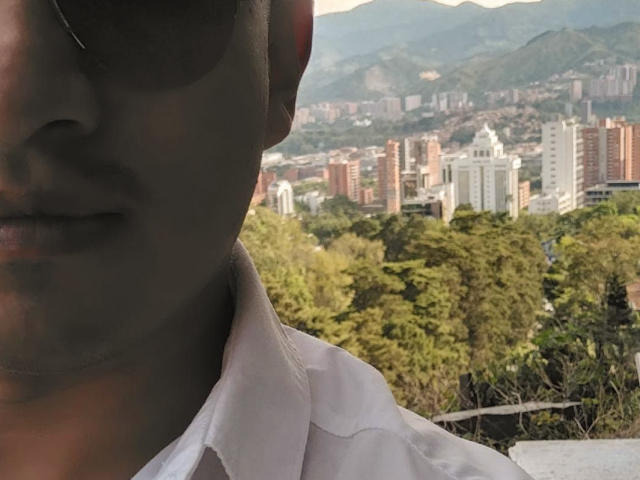 TerryHawttin's face, partial, in foreground, wearing white shirt, dark skin, sunglasses, mountain cityscape with buildings in background, green trees, urban and natural mix, slight shadow on face.