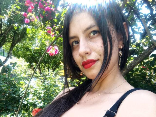 MadissonFontaine has red lipstick, black hair, and a cute face with small freckles, wearing a black strap top, and dangling earrings, standing in a sunny garden with pink flowers.
