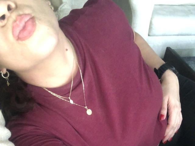 Yajayra's face, puffy pink lips, wearing a maroon shirt, gold necklace, earrings, black wristwatch, red nail polish, laying down, showing a small mole on her neck.