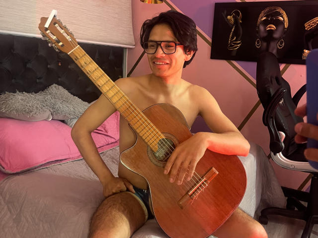 ThonixMr., shirtless with glasses, playing a wooden acoustic guitar, wearing blue briefs, smiling, sitting on a bed with pink pillows, and a black chair with a black mask.