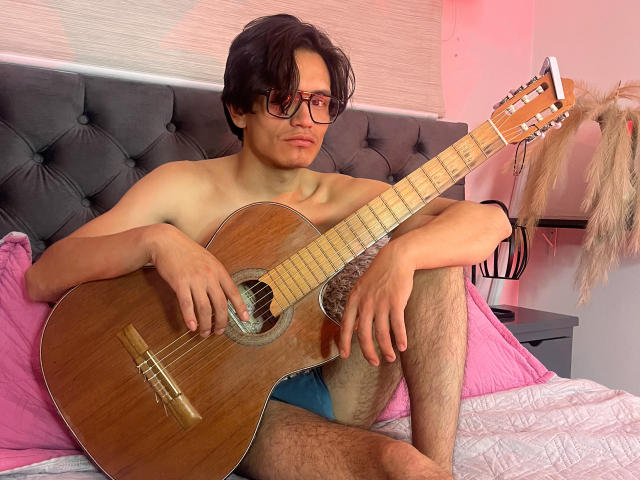 ThonixMr. with shaggy black hair, glasses, and a goatee, shirtless, holding a wooden acoustic guitar, sitting on a bed with pink pillows, wearing blue shorts.