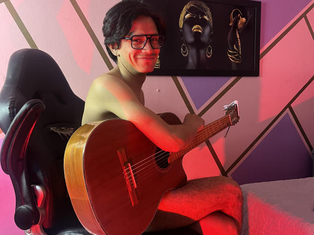 ThonixMr. is shirtless, with glasses, playing a wooden acoustic guitar while sitting on a black chair, smiling. Red and purple lighting highlights his hairy legs.