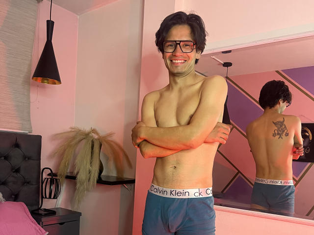 ThonixMr. stands shirtless with arms crossed, wearing blue Calvin Klein boxer briefs, smiling at the camera. He's got glasses, a cute face, and a tattoo on his back.