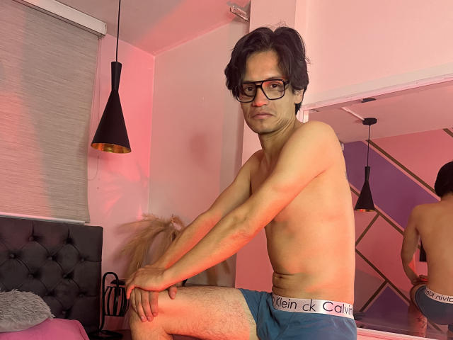ThonixMr. is a slim, shirtless dude with dark hair, glasses, and a serious face, sitting on a bed, wearing blue Calvin Klein underwear, showing his hairy legs.