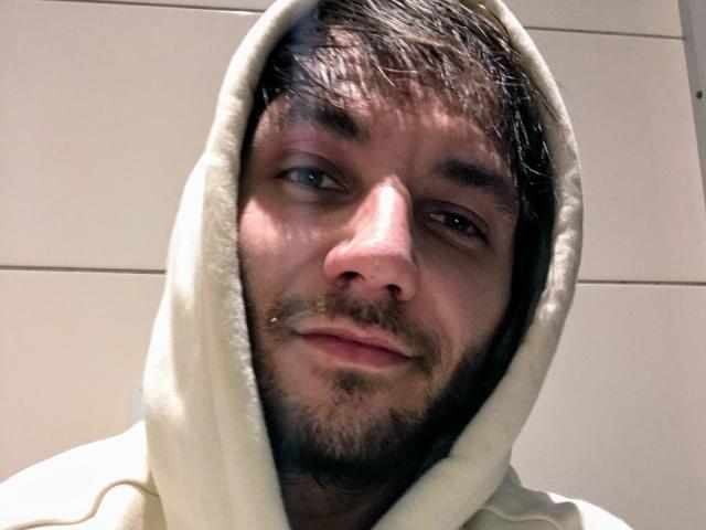 Le visage de JakieG, avec un léger sourire, porte un sweat à capuche blanc avec la capuche relevée. Il a les cheveux brun foncé, une barbe et un petit piercing au nez..