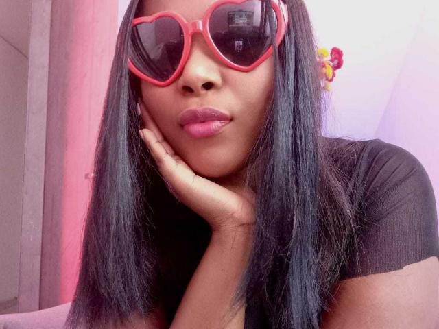 MacarenaBoobs with dark straight hair, pink heart-shaped sunglasses, and pink lipstick, resting her chin on her hand, wearing a black shirt, in a pink-lit room.