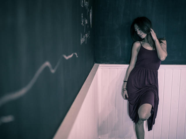 PoxyVibe has long dark hair, wearing a purple dress, standing with one leg raised, touching her head, white wainscoting, dark chalkboard wall, subtle smile, relaxed pose.