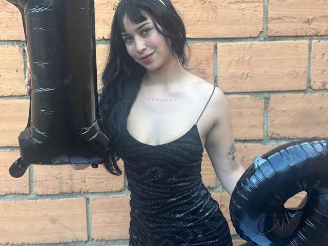 Sol Ortega, con cabello negro, vestida con un vestido negro brillante, sosteniendo globos negros, de pie frente a una pared de ladrillos, sonriendo ligeramente, mostrando pequeños senos y un tatuaje en su brazo..