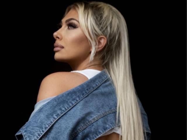 EmiliaFrench with long blonde ponytail, wearing a blue denim jacket over a white top, looking to the side with a serious expression, black background.