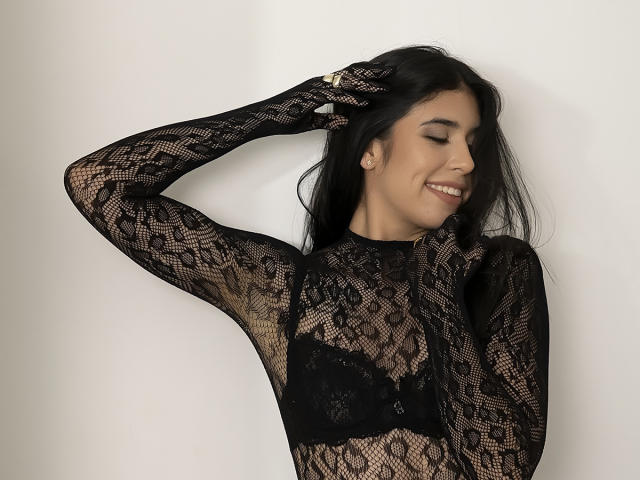MiluSky with black lace long-sleeve top, exposing her boobs in a black lace bra, smiling, eyes closed, right hand in hair, white background, ring on right hand.