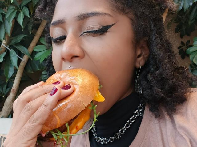MeganWlliams with curly hair eating a burger, her nose pierced, wearing black eyeliner and a chain necklace, her purple nails gripping the bun.