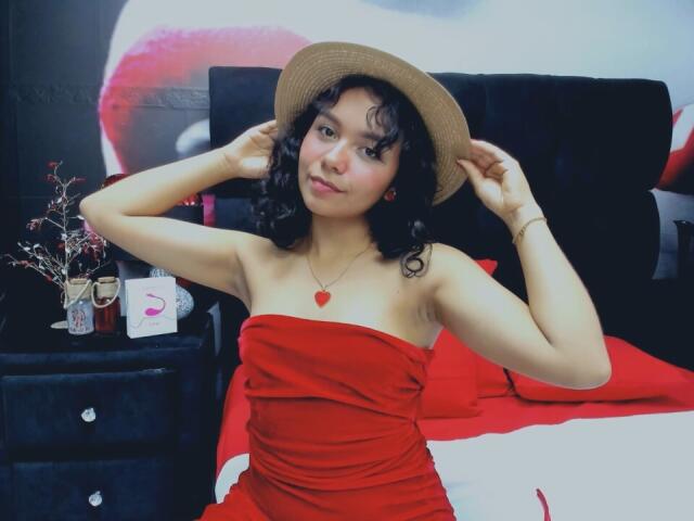 JuliaBennett with curly black hair, wearing a red strapless dress, red heart necklace, and a brown hat, sitting on a red and white bed, hands on hat.