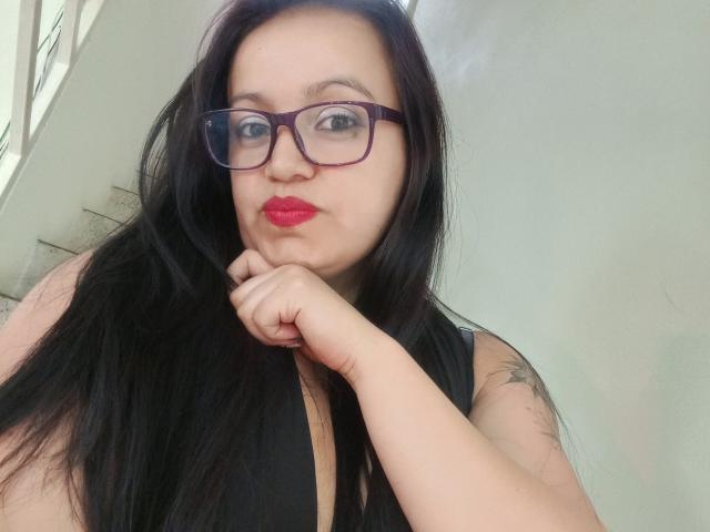 Ginebra Vargas mit langem, schwarzem Haar, rotem Lippenstift, Brille, schwarzem Top ohne Ärmel und einem Tattoo am rechten Arm, die die Lippen zusammenpresst und mit der Hand unter dem Kinn ruht, wobei sie ihre Brüste leicht zeigt..