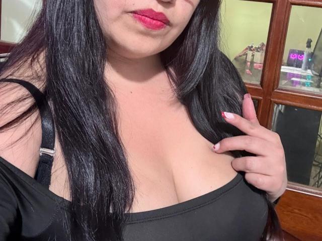 GinebraVargas with long black hair, red lipstick, and a black top, showing her big tits, one hand touching her chest. She has white nails and a wooden background.