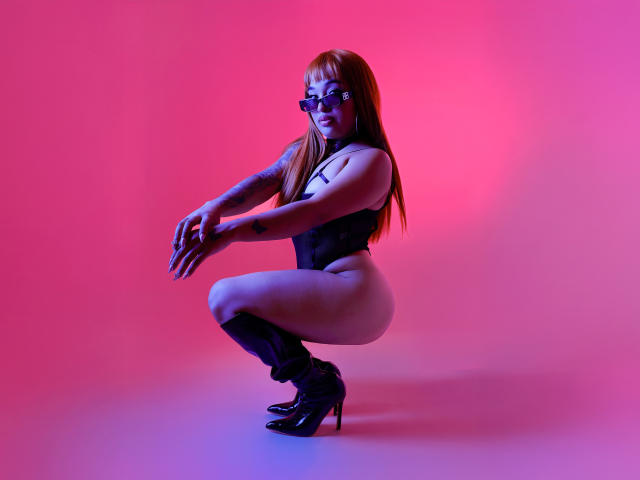 NikkiDean with long red hair, black glasses, and black high heels, squatting in black lingerie, showing her bare ass and tattooed arm, against pink and purple lighting.