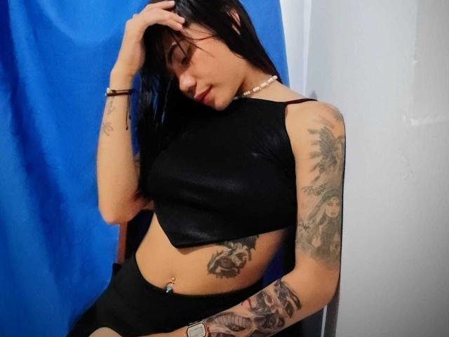 CristalleX trägt ein schwarzes Crop-Top und Shorts, hat Tattoos an Armen und an der Seite, schwarzes Haar, eine Hand im Haar, einen blauen Vorhang, ein Bauchnabelpiercing, trägt ein Armband und eine Halskette und sitzt..
