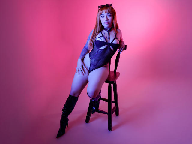 Nikki Dean mit roten Haaren, die einen schwarzen Badeanzug mit Ausschnitten, schwarze Stiefel und eine Halskette trägt, sitzt auf einem Stuhl, die Hände auf den Oberschenkeln, in rosafarbenem und blauem Licht. Brüste, Vulva, tätowierter Arm..
