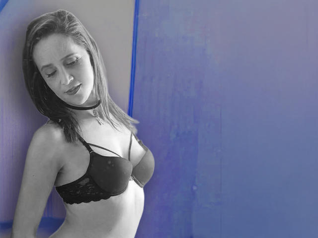 EmilyJunn with small tits, wearing a black lace bra and choker, has light skin and straight dark hair, standing against a blue wall, looking down with a subtle smile.