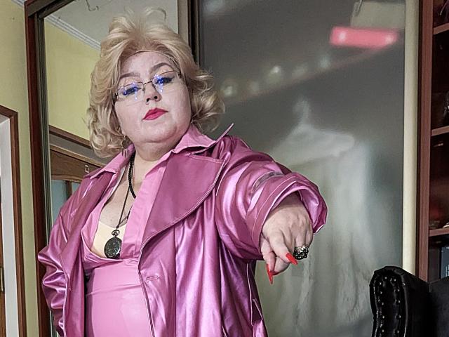 KaryQueen with curly blonde hair, blue eyeshadow, pink shiny jacket, and red nails, pointing forward, wearing a black pendant, in a room with a frosted glass door.