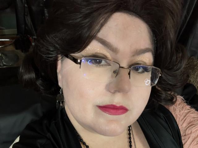 KaryQueen has white goo on her face, dark wavy hair, glasses, red lipstick, and is wearing a black top with a necklace. She's looking at the camera with a neutral expression.