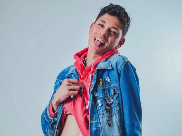 TylerCa, short dark hair, sticking out tongue, wearing blue denim jacket with pins, red hoodie, lifting shirt, showing flat stomach, light skin, slight smile, casual, cute.
