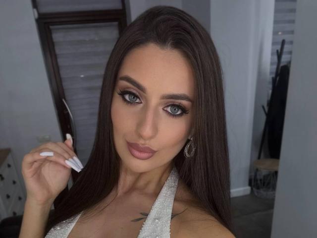 ArielGemme with long brown hair, green eyes, and white lace halter top, holding her white manicured nails, showing off her small tits and a tattoo on her chest.