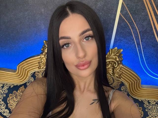 ArielGemme has long black hair, blue eyes, and full pink lips. She's wearing a gray top, showing a small black tattoo on her chest. Her tits are small.