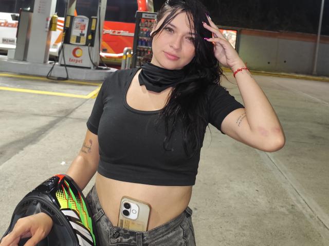 AlahiaStorm with black hair, wearing a black crop top and jeans, holding a black jacket, phone in front, tattoos on arms, mask around neck, night-time gas station.