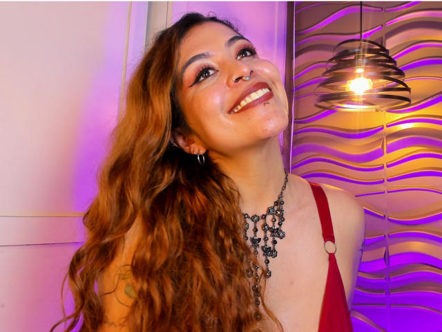 AlaiaLove with long wavy brown hair, smiling, wearing a red dress with a black necklace, nose ring, and hoop earrings, standing under a hanging light. Boobs partially visible.
