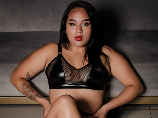 MillenaCollins with medium brown skin, black hair, and red lipstick, wearing a black mesh and leather bra, sitting with hands on knees, showing a tattoo on her right arm.