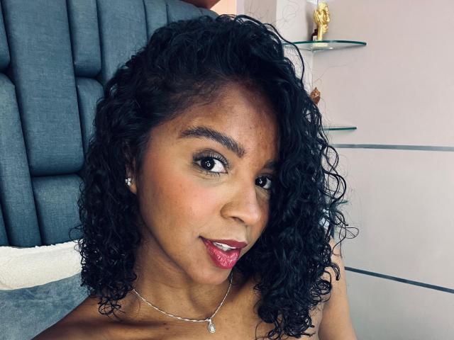 LisaMendez has dark curly hair, brown skin, and is smiling with pink lipstick. She's wearing a necklace with a small pendant and has diamond earrings.