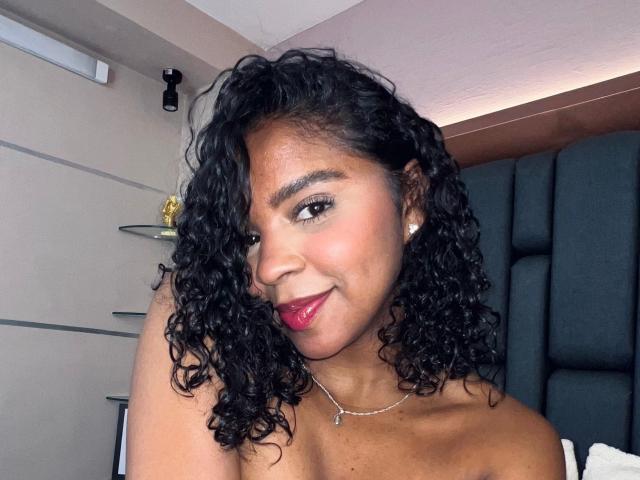 LisaMendez with curly black hair, medium brown skin, red lipstick, and a necklace, smiling at the camera, showing her bare shoulders and small, round, pearl earrings.