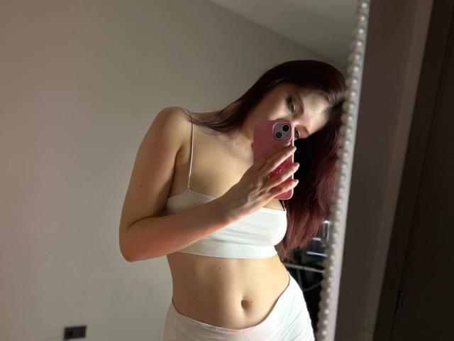 KessyMiu with red hair, holding pink phone, wearing white crop top, showing small tits and flat stomach, taking mirror selfie.
