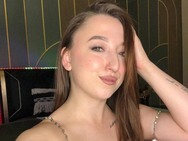 MonAmourR com traseiro volumoso, seios marrons e rosto brilhante, com um sorriso malicioso, a mão no cabelo castanho, olhos verdes, piercing no nariz e tatuagens, usando uma corrente de prata, em uma foto..