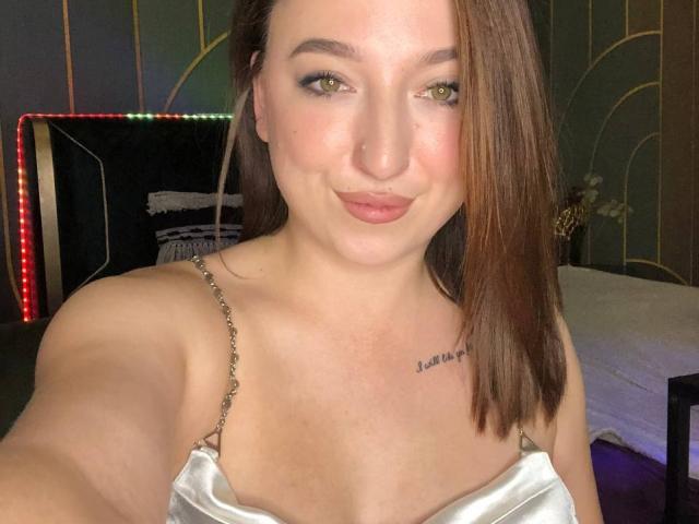 MonAmourR a la peau claire, les yeux verts et les cheveux bruns. Elle porte un haut blanc brillant avec des bretelles fines, laissant apparaître un petit tatouage sur sa poitrine, et elle a un sourire discret..