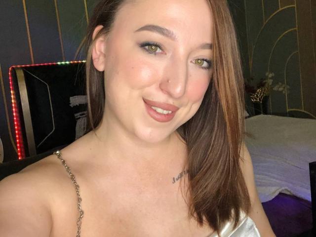 MonAmourR tem pele clara, olhos verdes e cabelo castanho, sorrindo com lábios rosados, mostrando uma pequena tatuagem no peito. Ela está vestindo um vestido com alças prateadas, e seus seios estão visíveis..