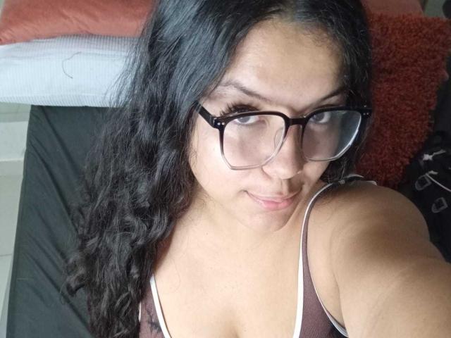 KonyBella, com longos cabelos pretos ondulados, usando óculos e uma camiseta marrom, mostrando seios pequenos e um rosto fofo, com um sorriso discreto, tirando uma selfie..