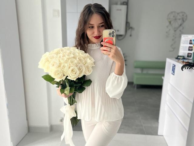 RozaSunn with white roses, wearing a white long-sleeve top and white pants, holding a bouquet in her right hand, taking a selfie with a red phone case.