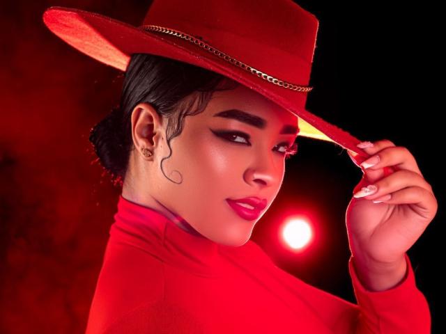 MilaRomano with dark hair in a bun, wearing a red hat and red turtleneck, holding the hat's brim, red lipstick, and gold earrings, against a dark background.