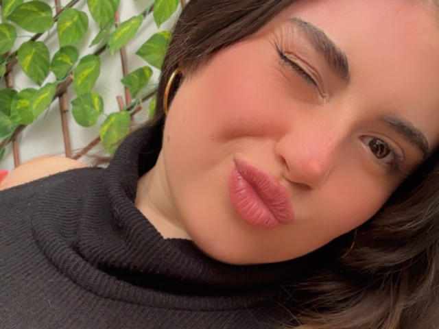 VicluX's face with a slight wink, pink pouty lips, short brown hair, black turtleneck, gold hoop earrings, and a light pink blush. Green leafy plant in the background.