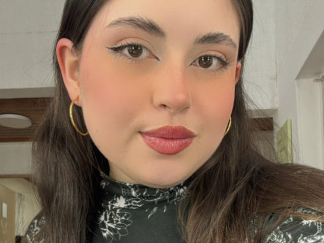 VicluX hat helle Haut, braune Augen, schwarzen Eyeliner, roten Lippenstift und langes, braunes Haar. Sie trägt goldene Ohrringe im Stecker-Stil und ein schwarzes Oberteil mit Blumenmuster. Ihr Hintern ist nicht sichtbar..