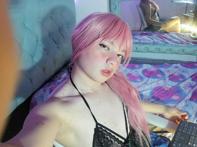 OliverPelman with pink hair, pouty lips, and black lace top, holding a keyboard, on a bed with a tufted headboard, visible butt tattoo. Reflective mirror in background.